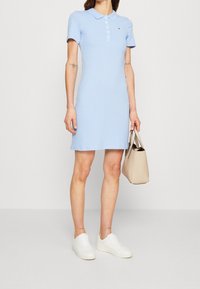 Lichtblauwe polojurk met een kraag, korte mouwen en een knoopsluiting. Gecombineerd met een beige handtas en witte sneakers.