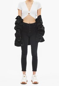 Weißes Crop-Top mit Cut-out-Detail, schwarze Jeansjacke, hochgeschnittene schwarze Jeans und weiße Sneaker mit braunen Sohlen.