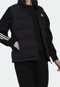 Svart pufferväst med hög krage, quiltat mönster, sidofickor och svart dragkedja. Vit Adidas-logotyp på bröstet, bärs över en svart topp.