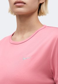 Κοντινό πλάνο ενός ατόμου που φοράει ροζ μπλούζα Nike με στρογγυλή λαιμόκοψη και μικρό ασημί λογότυπο Nike στο στήθος.