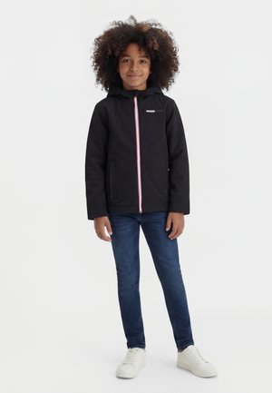 Göndör hajú gyermek fekete kapucnis dzsekit, kék farmert és fehér sportcipőt viselve, egyszerű fehér háttér előtt áll.