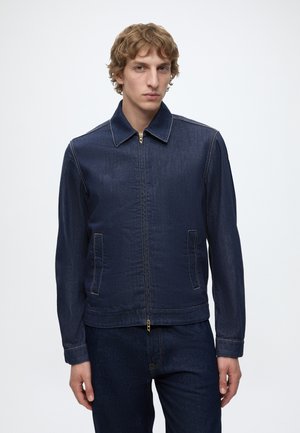 Junger Mann mit kurzen lockigen Haaren, der eine dunkelblaue Jeansjacke und passende Jeans vor einem schlichten hellen Hintergrund trägt.