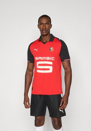 Un giovane si erge con sicurezza indossando una maglia da calcio rossa con maniche nere e pantaloni corti, calzini bianchi e un'espressione concentrata.