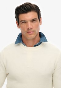 Biały sweter o teksturze z wysokim kołnierzem, nałożony na niebieską koszulę dżinsową. Kołnierz jest lekko widoczny w okolicy szyi.