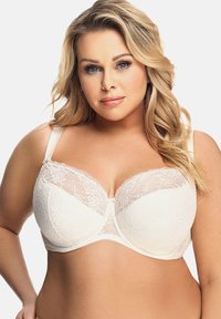 Mujer rubia con rizos sueltos que lleva puesto un sujetador de encaje blanco con aros, de frente y con una expresión neutral sobre un fondo liso.