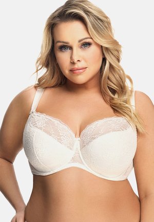 Mujer rubia con rizos sueltos que lleva puesto un sujetador de encaje blanco con aros, de frente y con una expresión neutral sobre un fondo liso.