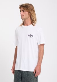 Homme aux cheveux mi-longs portant un t-shirt blanc à manches courtes avec un petit logo sombre sur la poitrine gauche et un pantalon foncé.