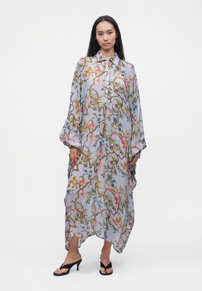 Femme portant une longue robe ample boutonnée avec un motif coloré de fleurs et de branches, associée à des sandales noires à talons hauts.