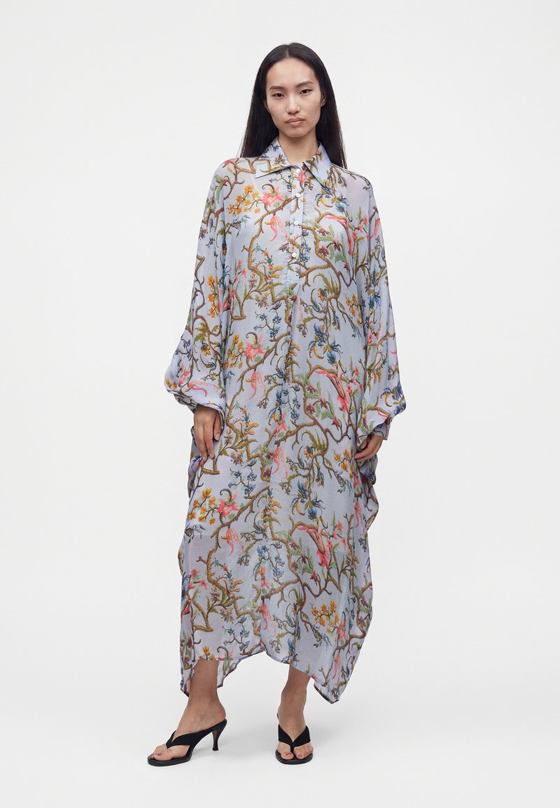 Femme portant une longue robe ample boutonnée avec un motif coloré de fleurs et de branches, associée à des sandales noires à talons hauts.