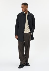 Muž v černém kabátu na knoflíky, béžovém svetru, hnědých širokých kalhotách a černých botách stojí s jednou rukou v kapse a dívá se doleva.