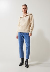 Pullover in pile beige con ampio colletto, chiusura a mezza zip, superficie texturizzata, polsini a costine, indossato con jeans in denim blu e stivaletti neri.