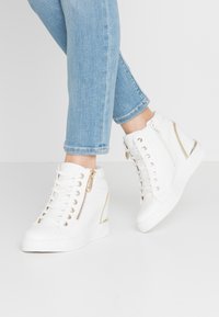 Person som bär ljusblå jeans och vita sneakers med hög skaft, med guld dragkedjor och präglad struktur, stående på vit golv.