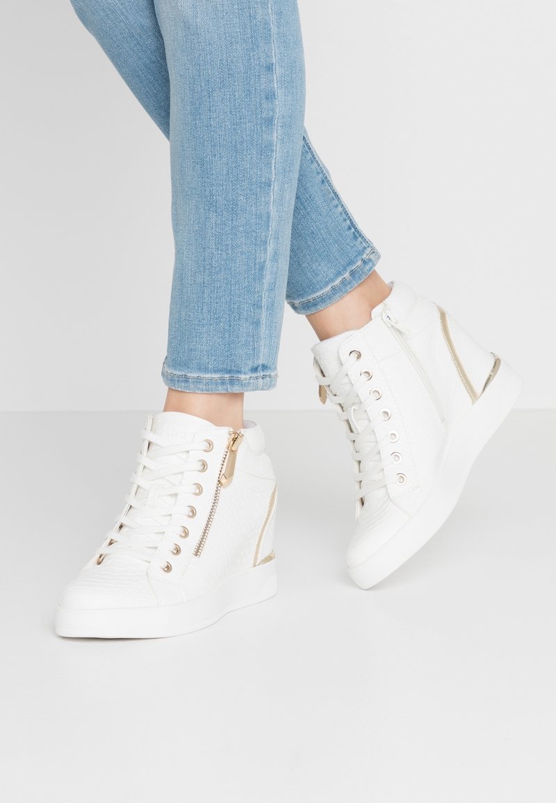 Person som bär ljusblå jeans och vita sneakers med hög skaft, med guld dragkedjor och präglad struktur, stående på vit golv.