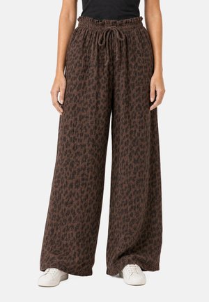 Weit geschnittene braune Hose mit dunklem Leopardenmuster, elastischem Kordelzugbund, getragen mit weißen Sneakers und schwarzem Oberteil, stehend vor einfachem Hintergrund.