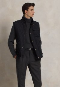 Chaleco negro acolchado con botones a presión y un bolsillo en el pecho, llevado sobre un suéter de cuello alto gris oscuro y pantalones grises. Textura suave, diseño minimalista.