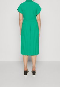 Vihreä laskostettu puolipitkä mekko lyhyillä hihoilla ja korkealla kauluksella, näkyy takaa päin. Litteät mustat korkokengät viimeistelevät asun.