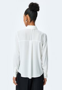 Blusa blanca con cuello, de manga larga y un ajuste ligeramente holgado. Tejido suave y ligero con un detalle de plisado en la parte posterior.