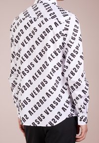 Witte shirt met een herhalend zwart "VERSUS" tekstpatroon. Lange mouwen met knoopmanchetten en een standaard kraag. Glad stofoppervlak.