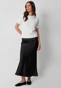 Biały, puszysty sweter z krótkim rękawem w parze z czarną satynową spódnicą maxi, z rozkloszowanym dołem i delikatnym połyskiem. Noszony z czarnymi płaskimi butami.