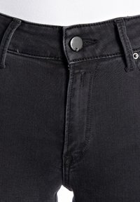 Primer plano de la cintura de unos jeans de mezclilla negra con cierre de botón, trabillas para cinturón y costura visible sobre un fondo blanco.