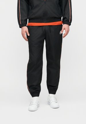 Homme portant un pantalon de survêtement noir avec des bandes latérales orange, un logo blanc, des baskets blanches et une veste zippée noire avec des détails orange.