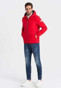 Giacca rossa con zip e cappuccio, dotata di tasche laterali e un tessuto testurizzato. Abbinata a jeans blu e sneaker bianche.