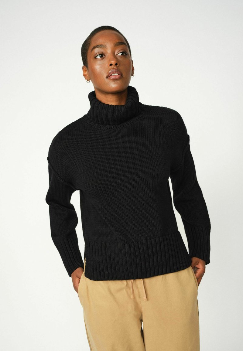 Maglione nero a maglia con colletto alto a coste e orlo largo a coste. Presenta spalle cadenti e un motivo testurizzato. Indossato con pantaloni beige.