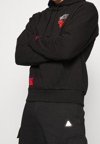 Felpa con cappuccio nera con logo e grafica rossa, dotata di polsini e orlo a coste. Abbinata a pantaloni della tuta neri con un accento triangolare.