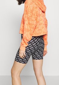 Orange kortärmad huvtröja med böljande mönster, kombinerad med svarta shorts som har upprepade vita "VANS"-logotyper och rutiga detaljer.