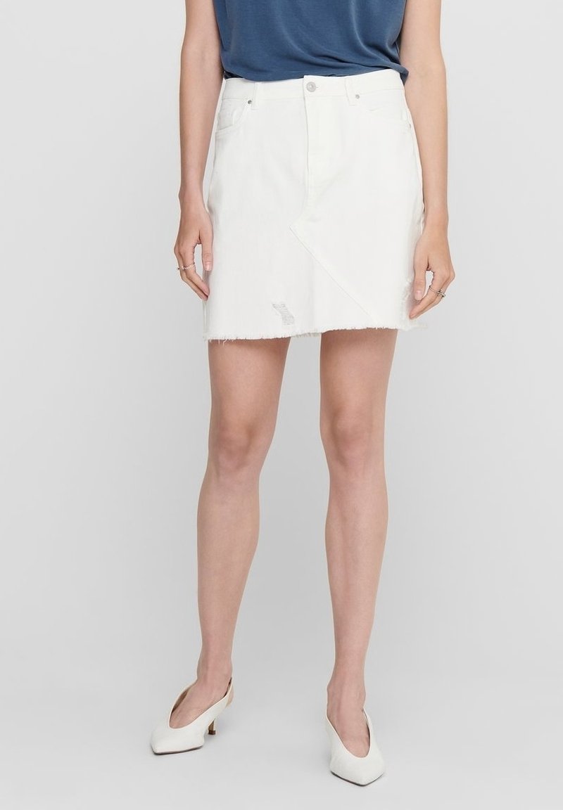 Jupe en denim blanc avec un ourlet effiloché, un design asymétrique, des détails légèrement usés et une fermeture à boutons à l'avant. Associée à des talons blancs.