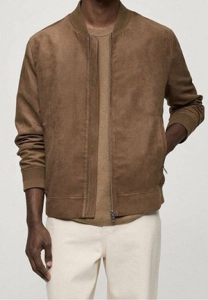 Homme portant une veste bomber en daim marron sur un pull beige et un pantalon blanc cassé, une main dans la poche de la veste.