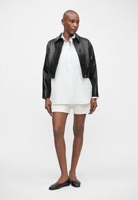 Μαύρο κοντό δερμάτινο τζάκετ, λευκή πουκαμίσα, μπεζ σορτς και μαύρα φλατ παπούτσια που φοράει ένα μοντέλο. Λείες υφές και μινιμαλιστικός σχεδιασμός.