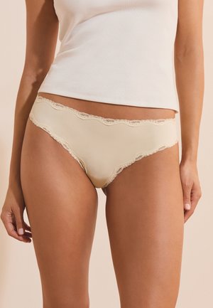 Kobieta ubrana w beżową bieliznę w stylu bikini ozdobioną koronką oraz biały prążkowany top na ramiączkach, stojąca na jednolitym tle.