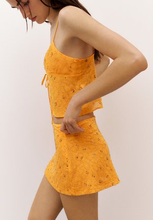 Femme portant une tenue orange ornée en deux pièces avec un haut sans manches et une mini-jupe ajustée sur un fond uni.