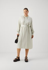 Robe midi rayée vert clair avec une ceinture à nouer, des manches longues et des poignets élastiques ; accompagnée de sandales à plateforme noires et d'un petit sac à main.