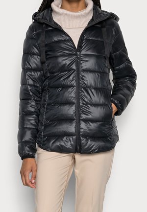 Personne portant une doudoune noire brillante avec capuche, un pull à col roulé beige et un pantalon beige clair, debout avec une main dans une poche.