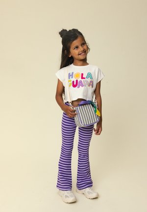 Lächendes Mädchen, das ein weißes Crop-Top mit der Aufschrift "HOLA GUAPA", lila gestreifte Hosen, weiße Sneakers trägt und eine kleine gestreifte Umhängetasche dabei hat.