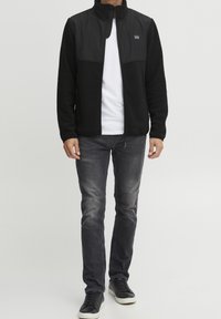 Mand iført en sort zip-up jakke over hvid skjorte, grå slidte jeans og sorte sneakers med hvide såler, stående mod en ensfarvet baggrund.