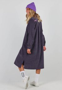 Robe longue en velours côtelé violet foncé avec une coupe décontractée, fentes latérales et poignets boutonnés, associée à un bonnet violet et des baskets blanches.