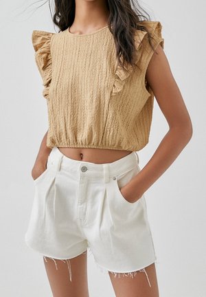 Kobieta ubrana w beżowy, bezrękawnik z teksturowanego materiału, z falbankami na ramionach, oraz białe, wysokiej talii szorty z dżinsu z postrzępionymi brzegami.