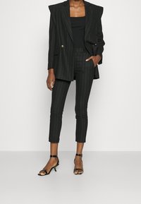Black pinstriped suit featuring a structured blazer with padded shoulders and tapered trousers. Worn with a fitted black top and strappy heels.