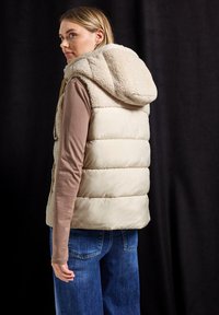 Gilet matelassé beige avec une capuche duveteuse, présentant une texture matelassée et un corps en tissu lisse, porté sur un haut marron à manches longues et un jean.