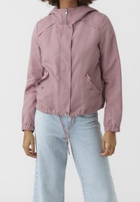 Kvinna iklädd en mauve zip-up jacka med sidofickor och ljusblå högmidjade jeans, stående mot en vit bakgrund.