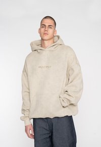 Beige Übergröße Hoodie mit einer vorderen Tasche und Kapuze mit Kordelzug. Verfügt über einen strukturierten Stoff und ein dezentes "MULTIPLY"-Logo auf der Brust.