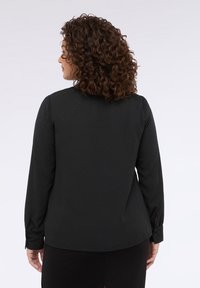 Blusa nera a maniche lunghe con una leggera texture a motivo, polsini con bottoni e orlo arrotondato, caratterizzata da un tallone posteriore per una vestibilità strutturata.