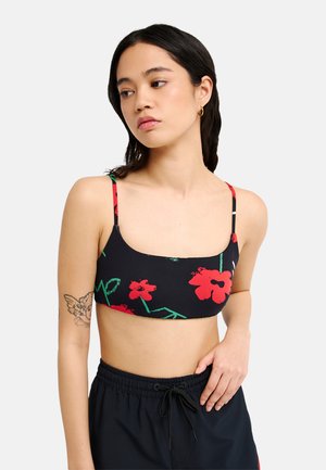 Junge Frau trägt ein schwarzes, geblümtes Crop-Top und schwarze Hose mit Kordelzug, mit einem Tattoo am Oberarm, schaut zur Seite.