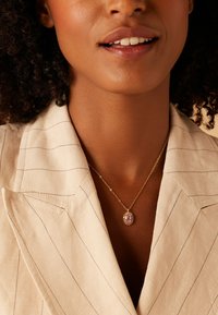 Gouden hangerketting met een ovale roze edelsteen en een halo-ontwerp, rustend op een delicate ketting tegen een beige gestreepte blazer.