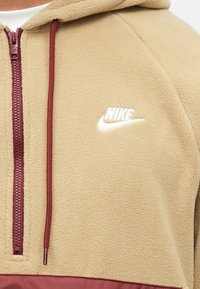 Hnedý fleecový pulóver s marhuľovým zipsom a akcentmi. Obsahuje biele logo Nike na hrudi a textúrovanú látku.