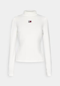 White long-sleeve turtleneck shirt made of soft fabric, featuring a small, branded logo patch in red, white, and blue on the chest.