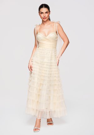 Mujer con cabello oscuro lleva un vestido de tul marfil sin mangas, con volantes y lazos en los hombros, y tacones blancos abiertos en los dedos, de pie frente a un fondo liso.